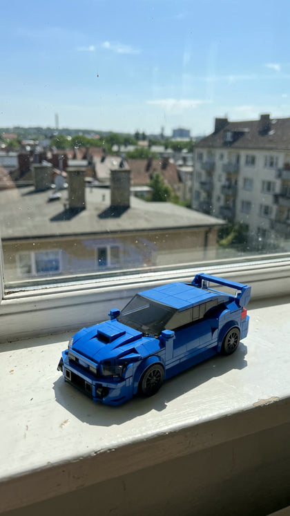 A blue brick-built rally saloon on a sunny windowsill with a city view